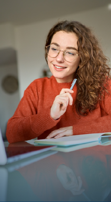 Close-up of a smiling woman watching something on her laptop while taking notes in a notebook. 
